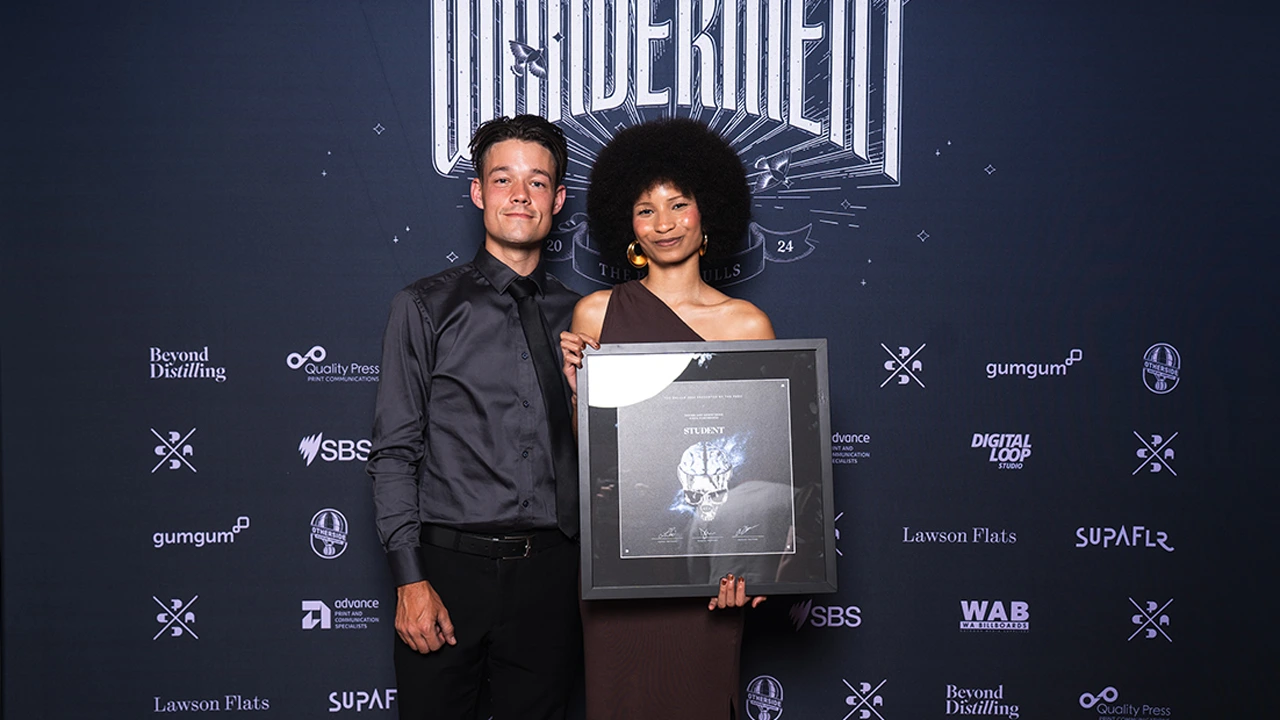 A person holding a framed award at a Perth Advertising and Design Club event, standing in front of a branded backdrop with sponsor logos