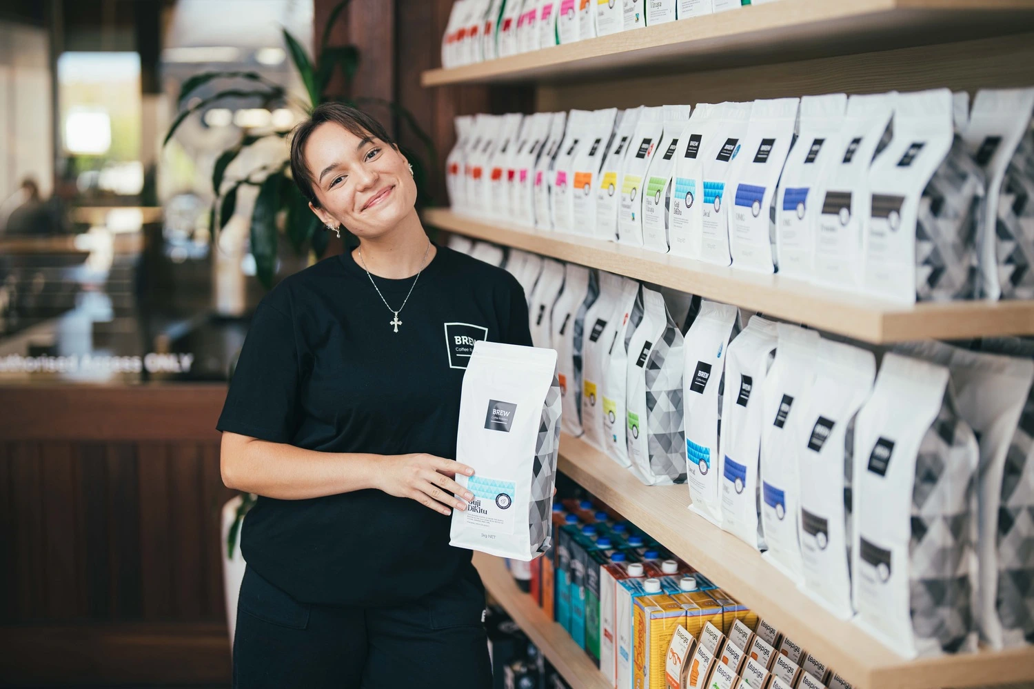 A woman standing behind a coffee counter with rows of Brew Coffee branded bags visible on shelves behind her