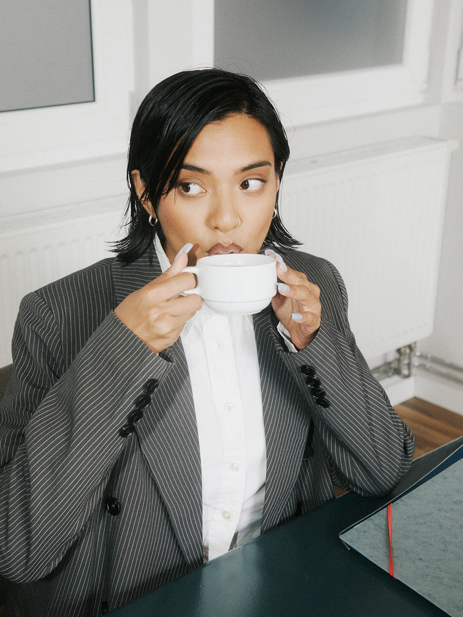A woman in a grey blazer sipping from a coffee cup against a dark background — Brew Coffee lifestyle portrait