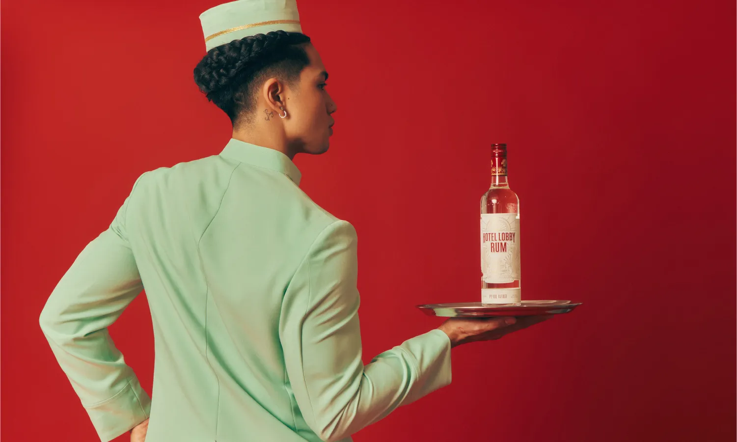 Woman in mint green hotel uniform holding a serving tray with Hotel Lobby Rum bottle on red background