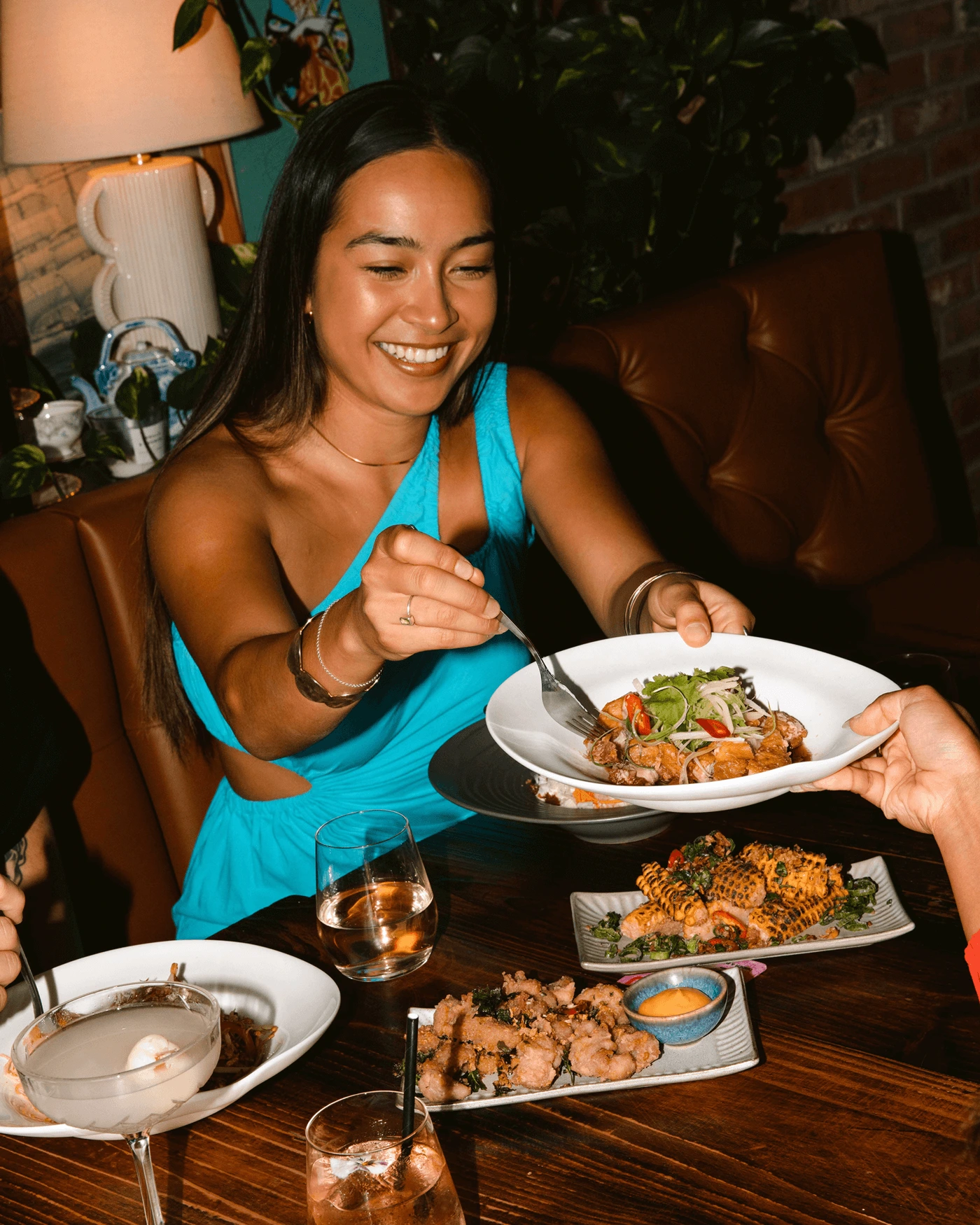 A young woman in a bright blue top smiling while holding a food dish in a restaurant setting — Lucy Lou campaign photography