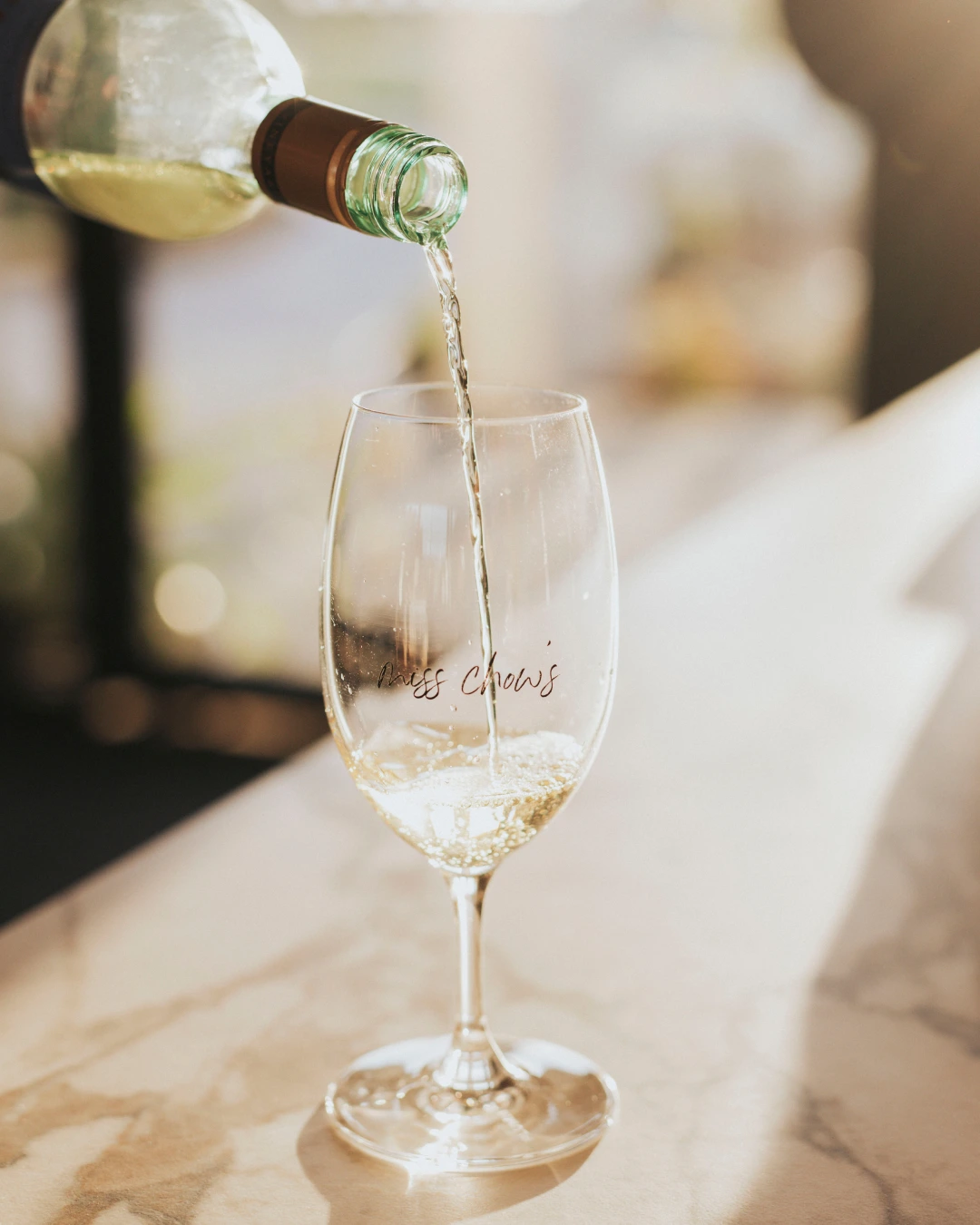 Close-up of white wine or champagne being poured into a glass with a soft bokeh background — Miss Chow's Group beverage photography
