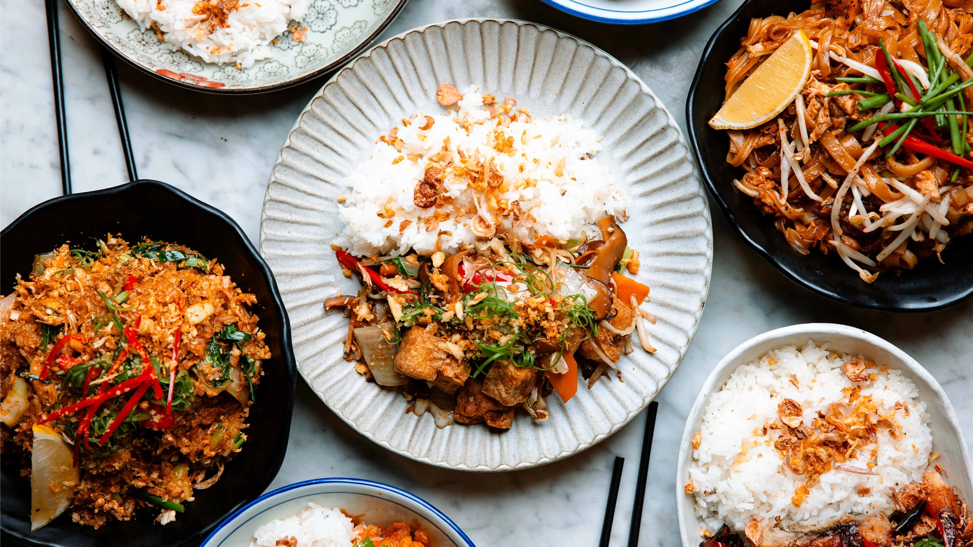 Overhead food photography showing a spread of vibrant Asian dishes including stir-fries, rice and chopsticks on a table