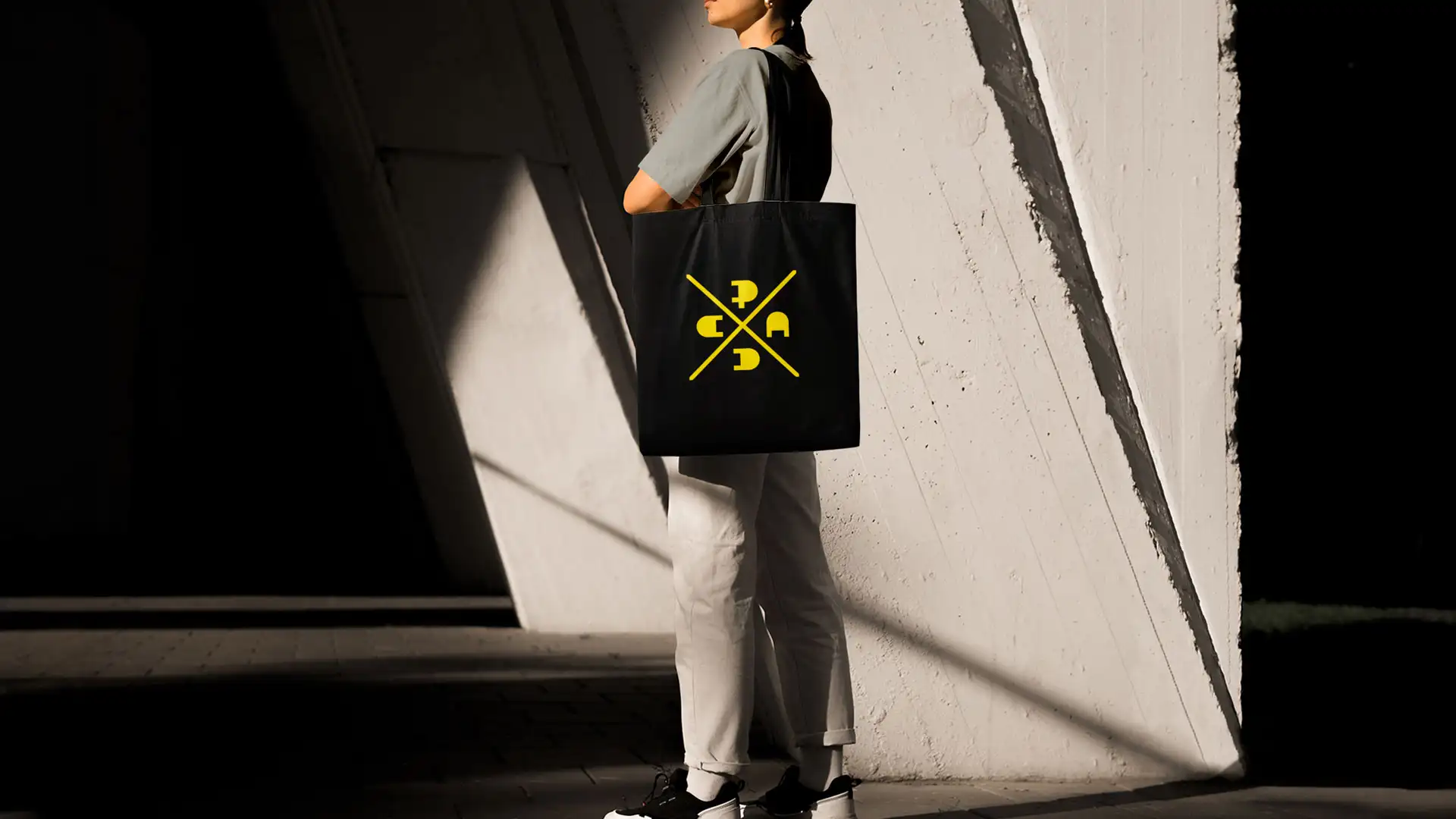 Person holding a black tote bag with a gold PADC brand design against a bright white sunlit background