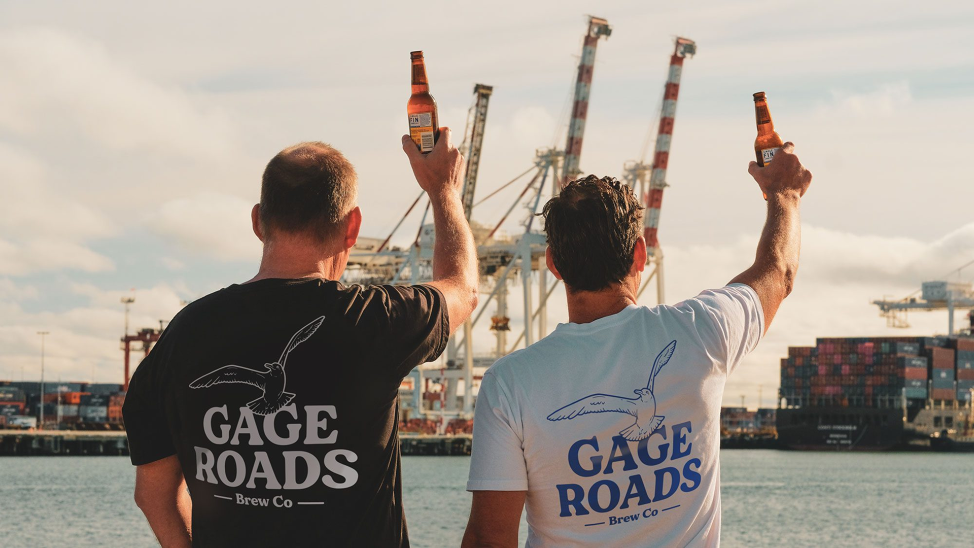 Two men in Gage Roads branded t-shirts raising beer bottles toward the harbour from behind