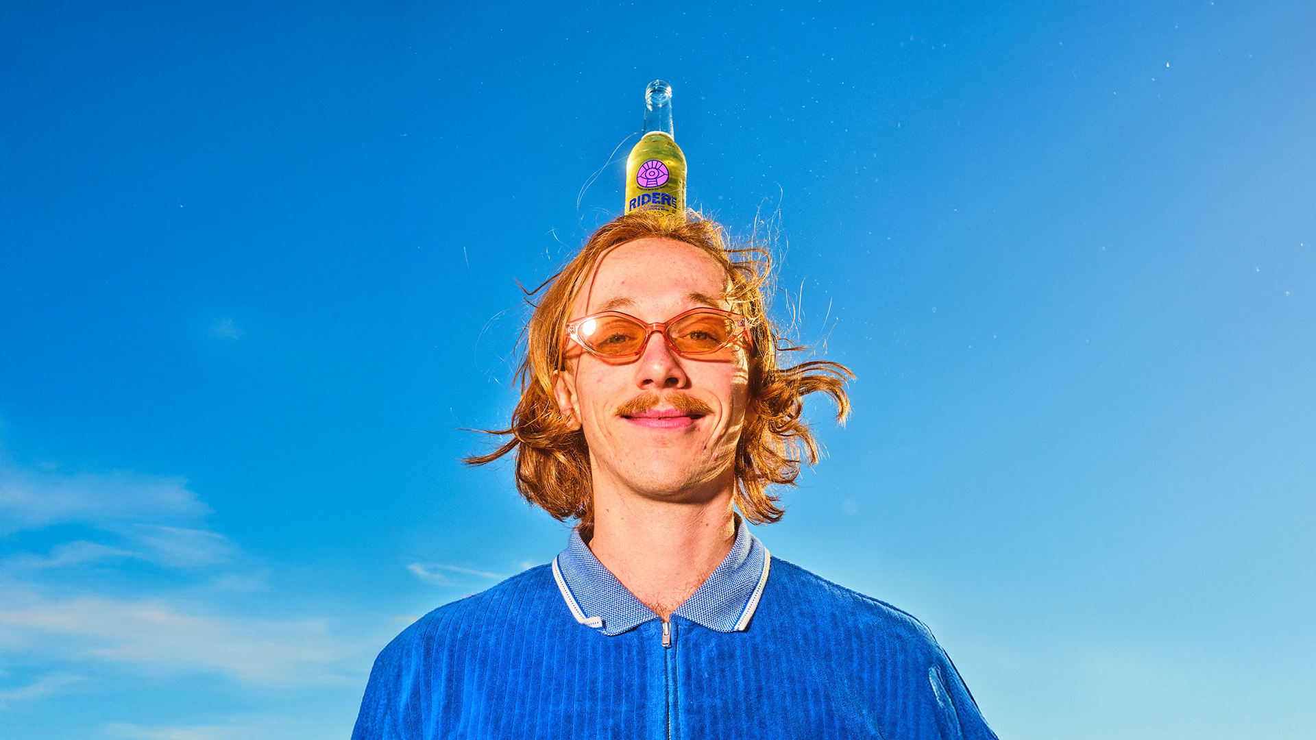 Young man with curly blonde hair and sunglasses balancing a bottle on his head at the beach
