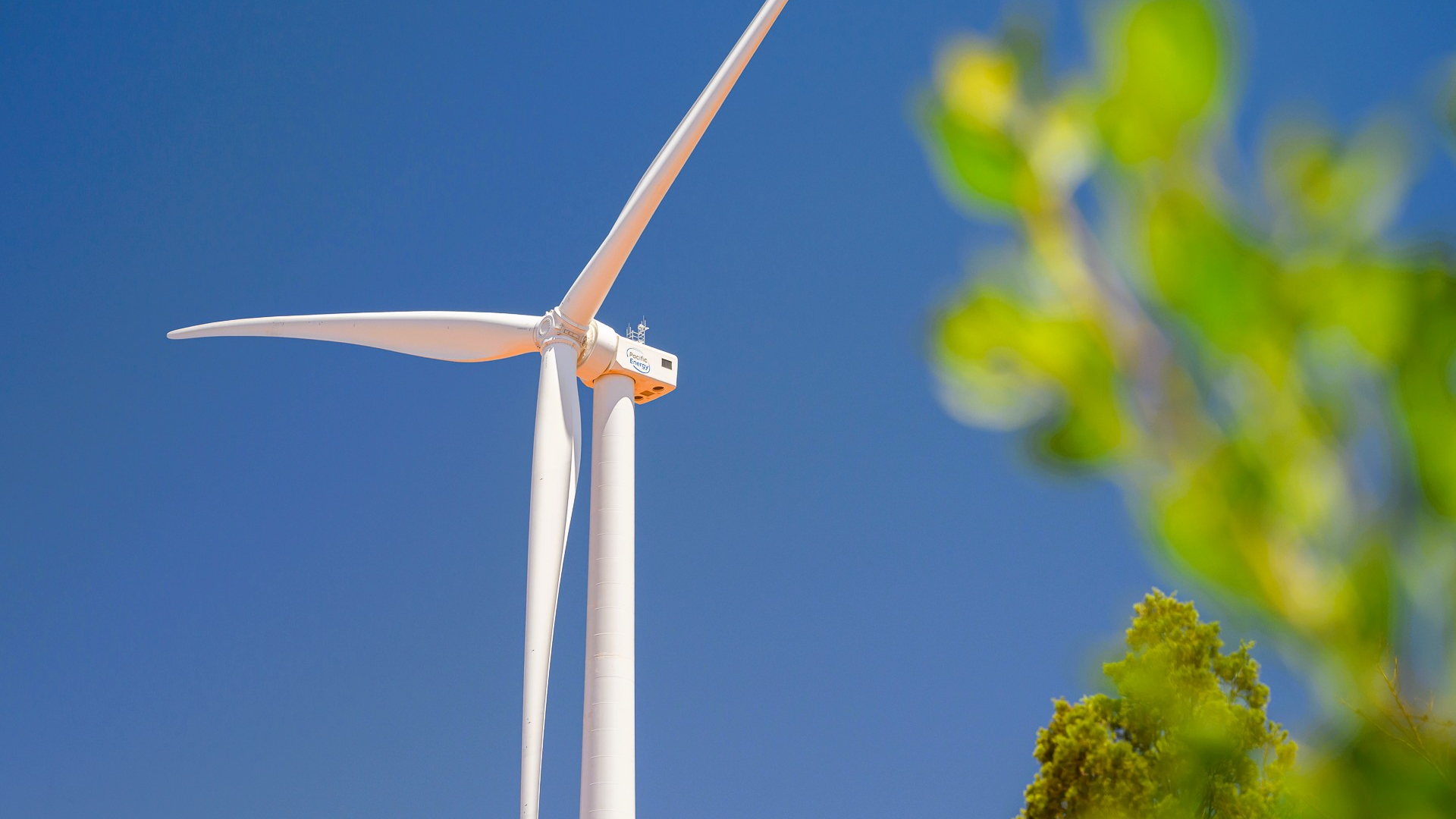 Wind turbine against a clear blue sky with green trees in the foreground
