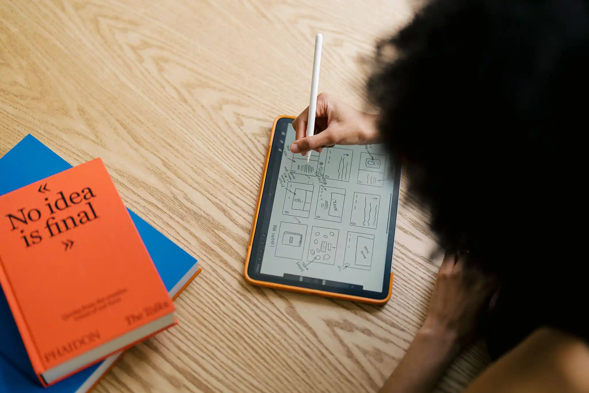 Top-down view of a designer's hands using a stylus on a tablet, surrounded by colourful design reference cards and books